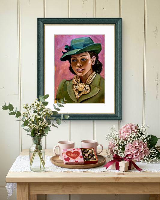 A framed art print featuring a colorful portrait of a woman in a green hat and coat, displayed on a light wood table with flowers, mugs, and a gift box against a white paneled wall.